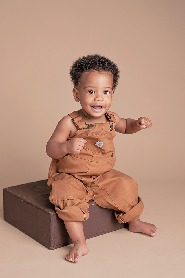 Smiling baby boy in brown Carhartt overalls sitting on a dark box against a tan studio background.