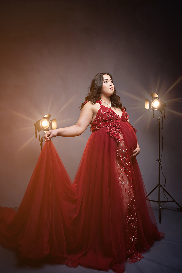 Pregnant woman in elegant red sequin gown with tulle train poses gracefully in studio with dramatic lighting.