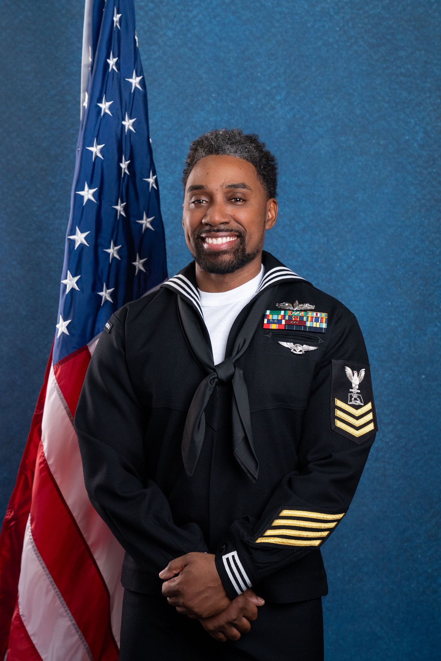 Smiling Navy sailor in dress uniform with medals and gold chevrons poses before an American flag on a blue background.