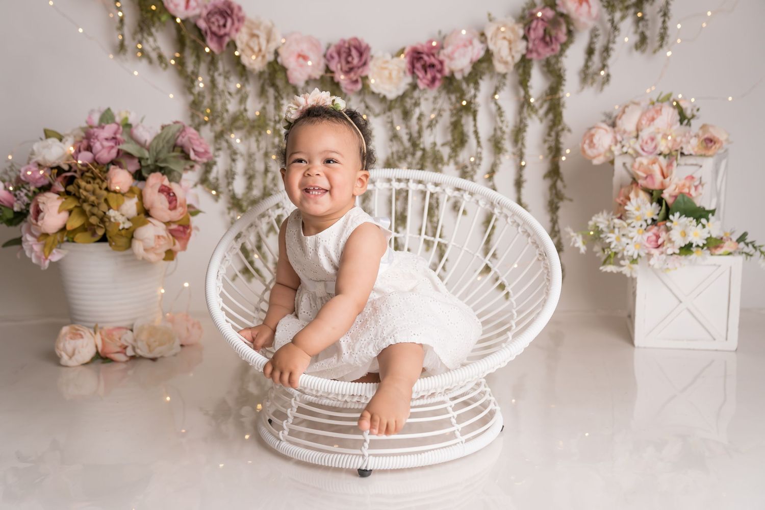 Baby in white dress sitting in wicker peacock chair with floral and pink balloon backdrop. San Diego cake smash photographer
