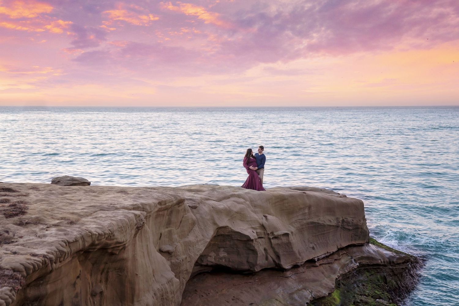 Romantic coastal photoshoot on rocky cliffs overlooking ocean at sunset with pink and purple sky. San Diego Maternity Photographer