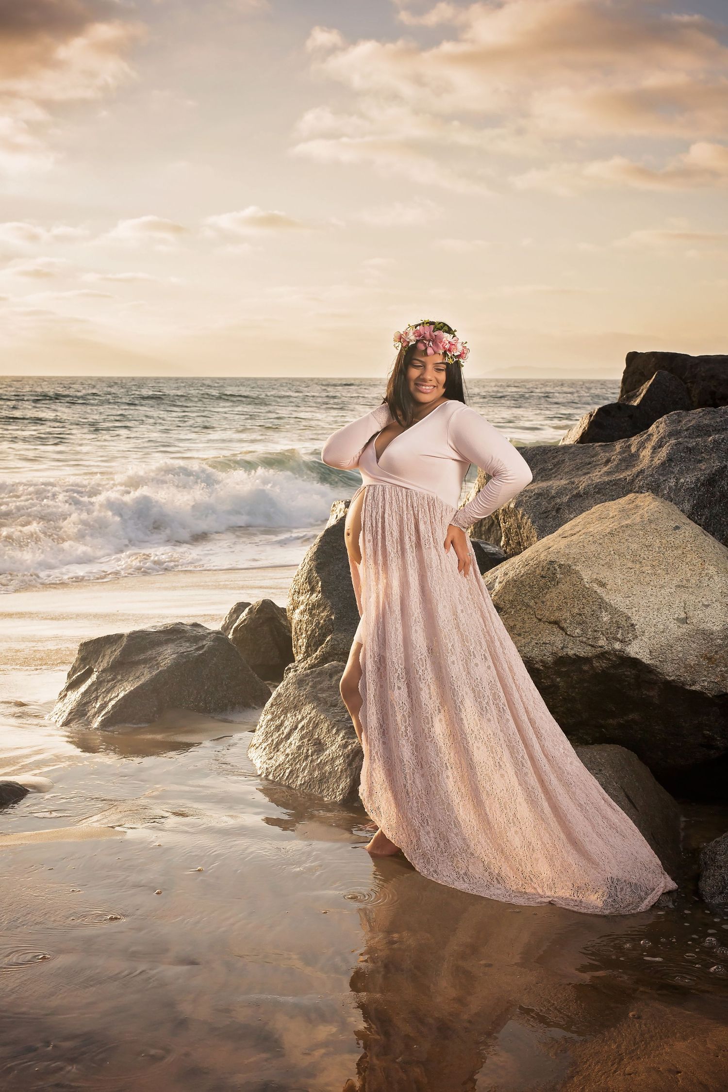 Pregnant woman in flowing pink dress poses on rocky beach during golden hour sunset. San Diego Maternity Photographer