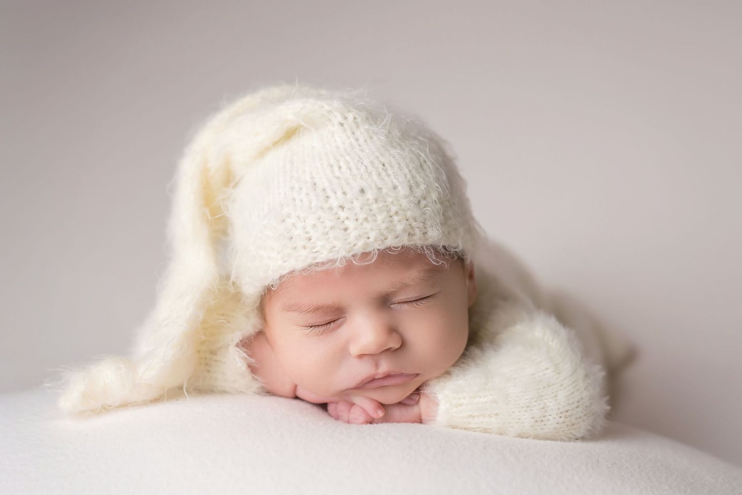 Sleeping baby wearing white knitted hat resting on soft white surface in san diego newborn photography studio.