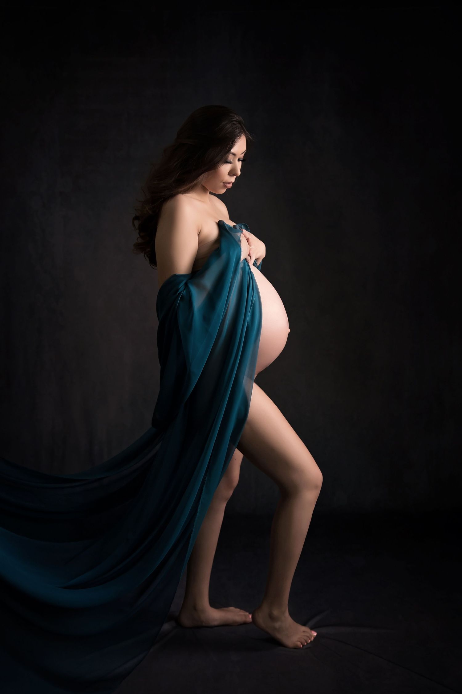 Artistic silhouette of expectant figure draped in teal fabric against dark background.