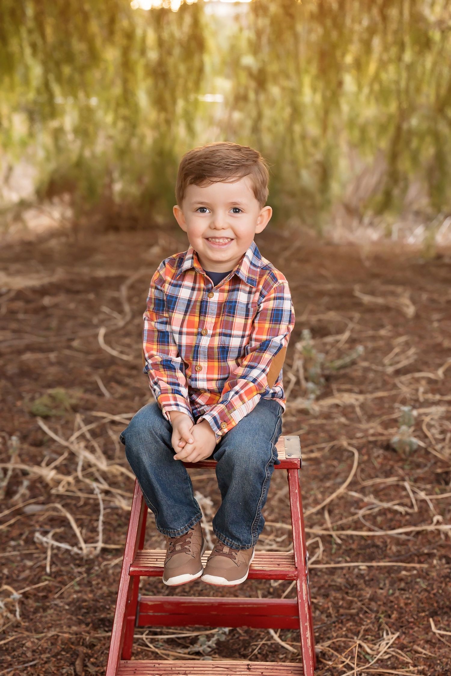 Young boy in plaid shirt smiling while sitting on a red stool in a natural outdoor setting