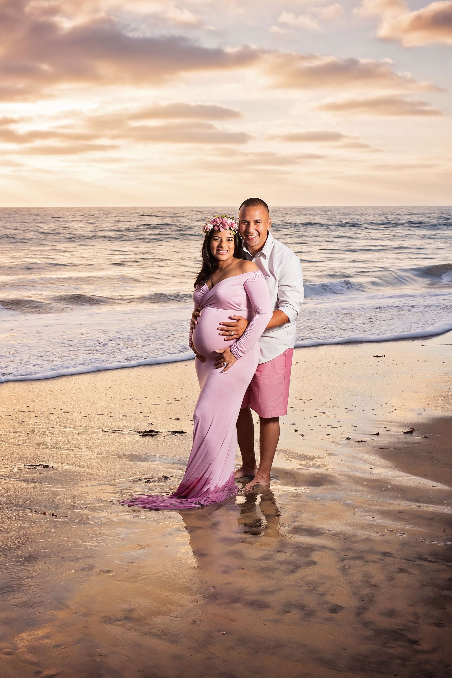 Sunset beach maternity photoshoot with couple in pink attire standing in ocean waves. San Diego Maternity Photographer