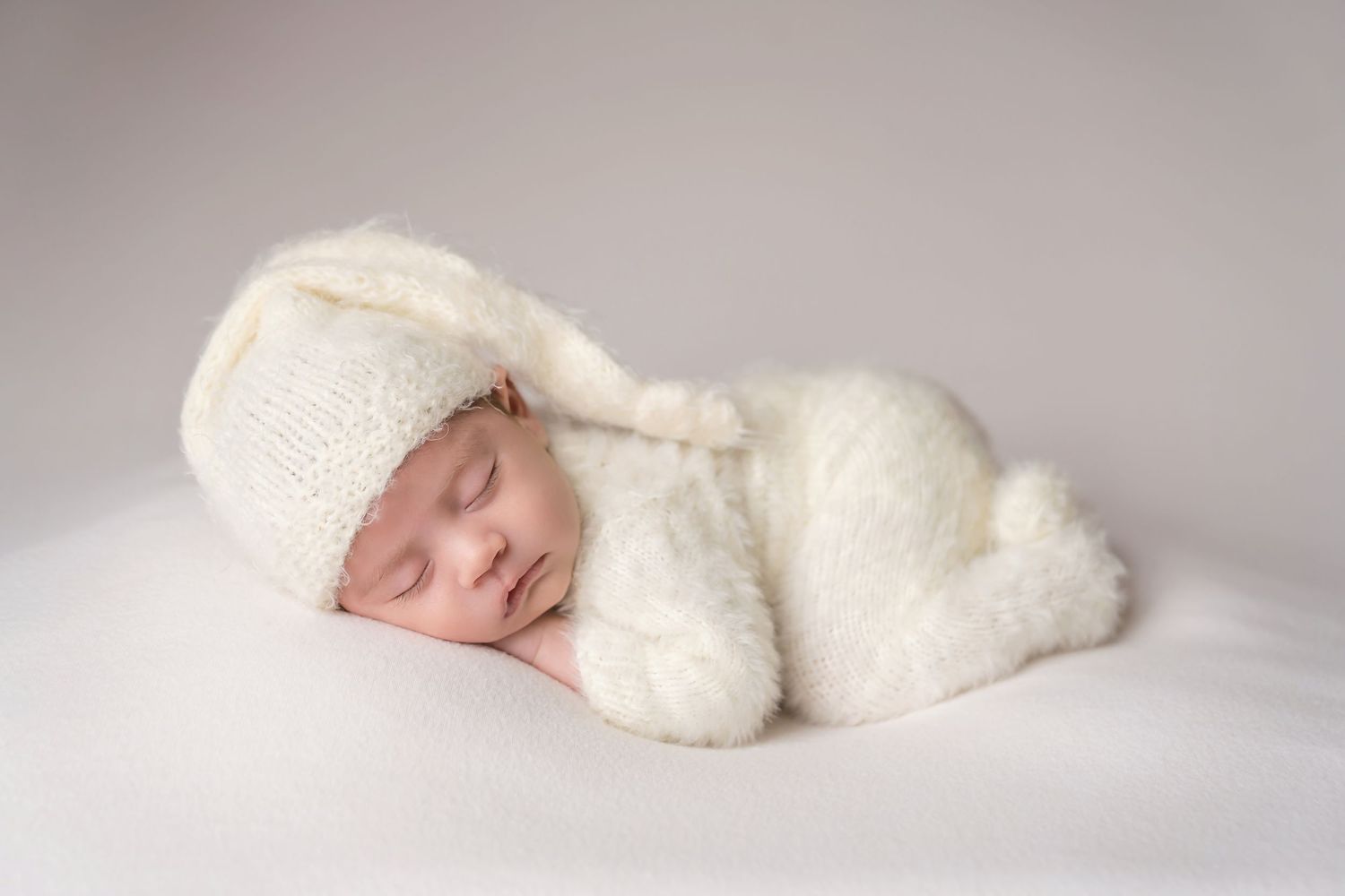 Peaceful sleeping baby wearing cozy white knit outfit on soft white backdrop. San Diego Newborn Photographer