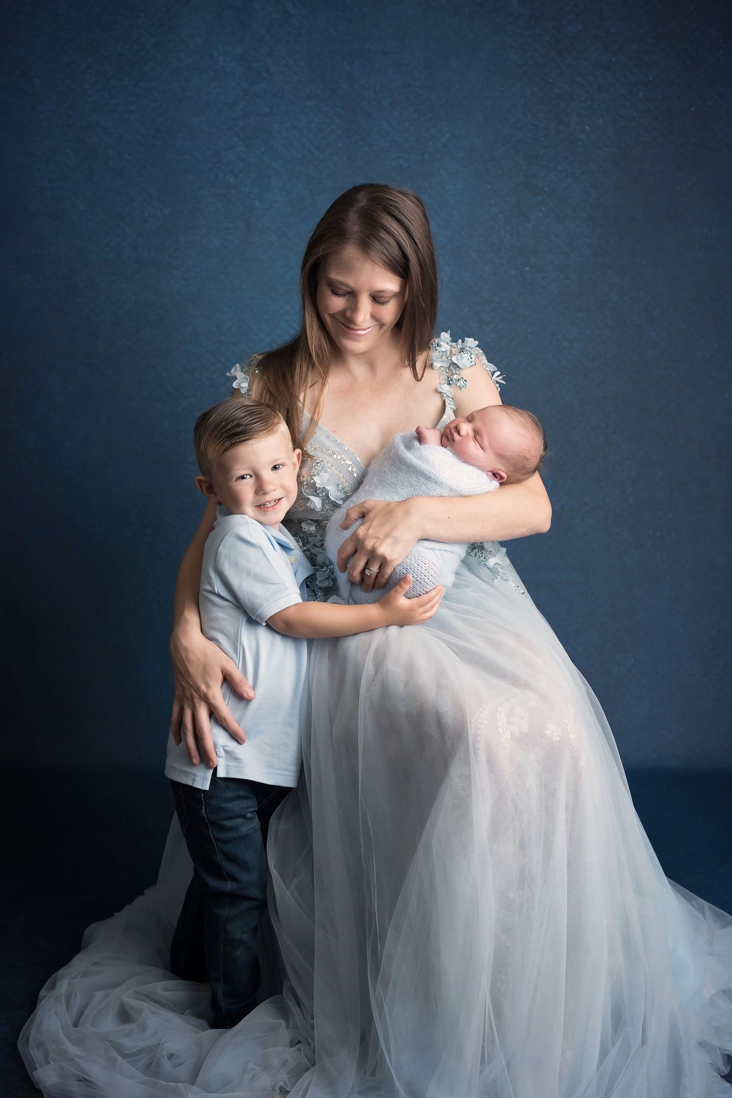 Family portrait with flowing white dress against deep blue studio backdrop. San Diego Newborn Photographer