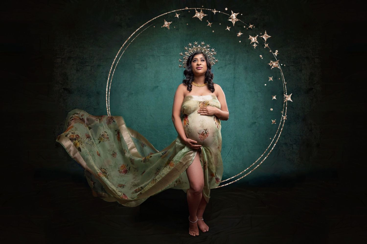 Pregnant woman in flowing green gown and star crown poses gracefully inside a glowing celestial hoop against teal backdrop.