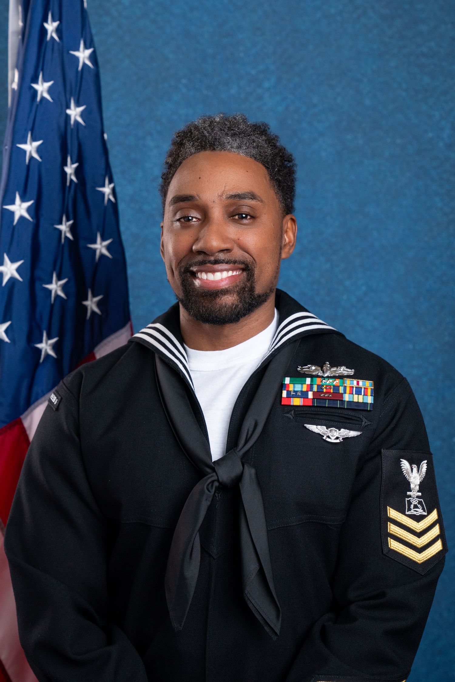 US Navy sailor in dress uniform smiling before American flag, displaying service ribbons and rank insignia.