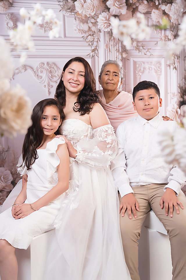 Family portrait of four generations in white outfits posing before floral backdrop in elegant studio setting.