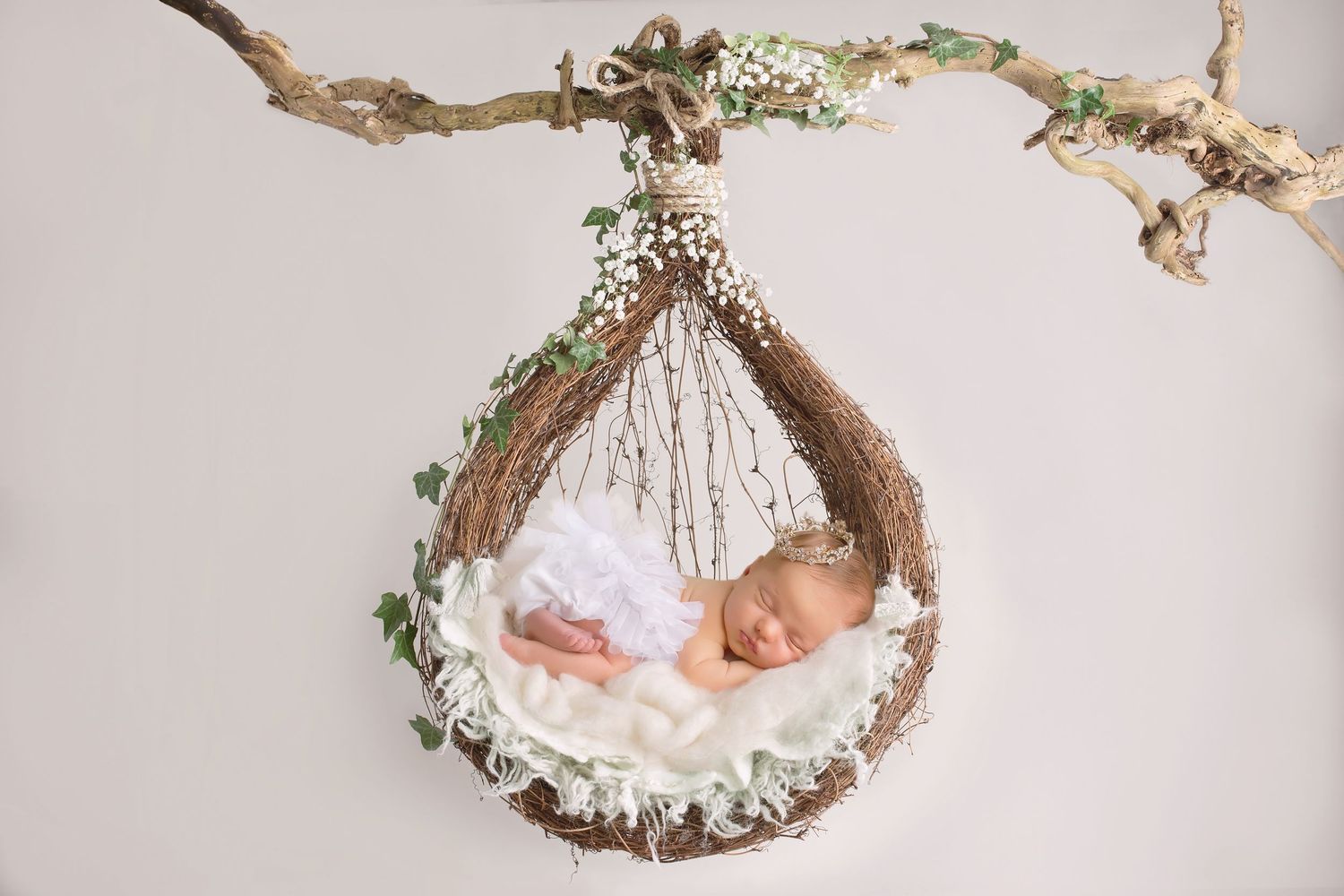 Newborn baby sleeping in suspended nest decorated with delicate flowers and greenery. San Diego Newborn Photographer