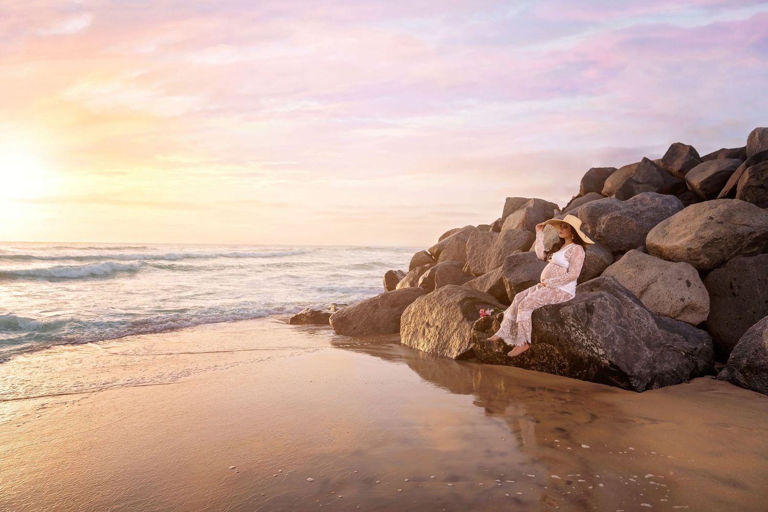Maternity photo in light pink dress contrasts against coastal rocks during golden hour maternity photography in san diego beach .
