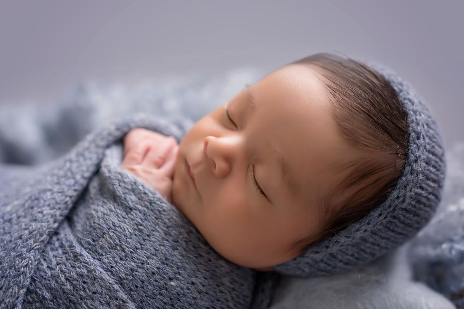 Sleeping newborn in gray knit wrap against dark background in artistic portrait. San Diego Newborn Photographer