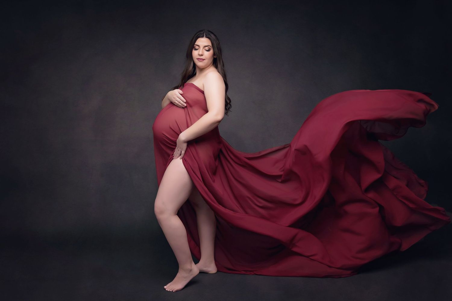 Maternity portrait with flowing burgundy dress against a deep studio backdrop