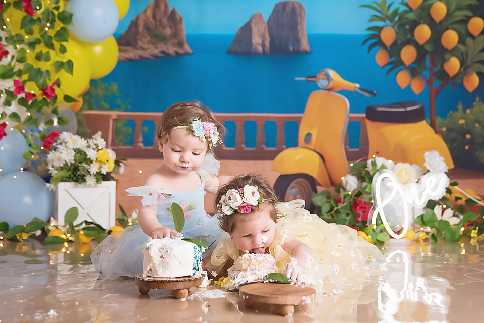 Two baby girls in floral headbands enjoy cake smash session with colorful Italian-themed backdrop featuring scooter and lemons.