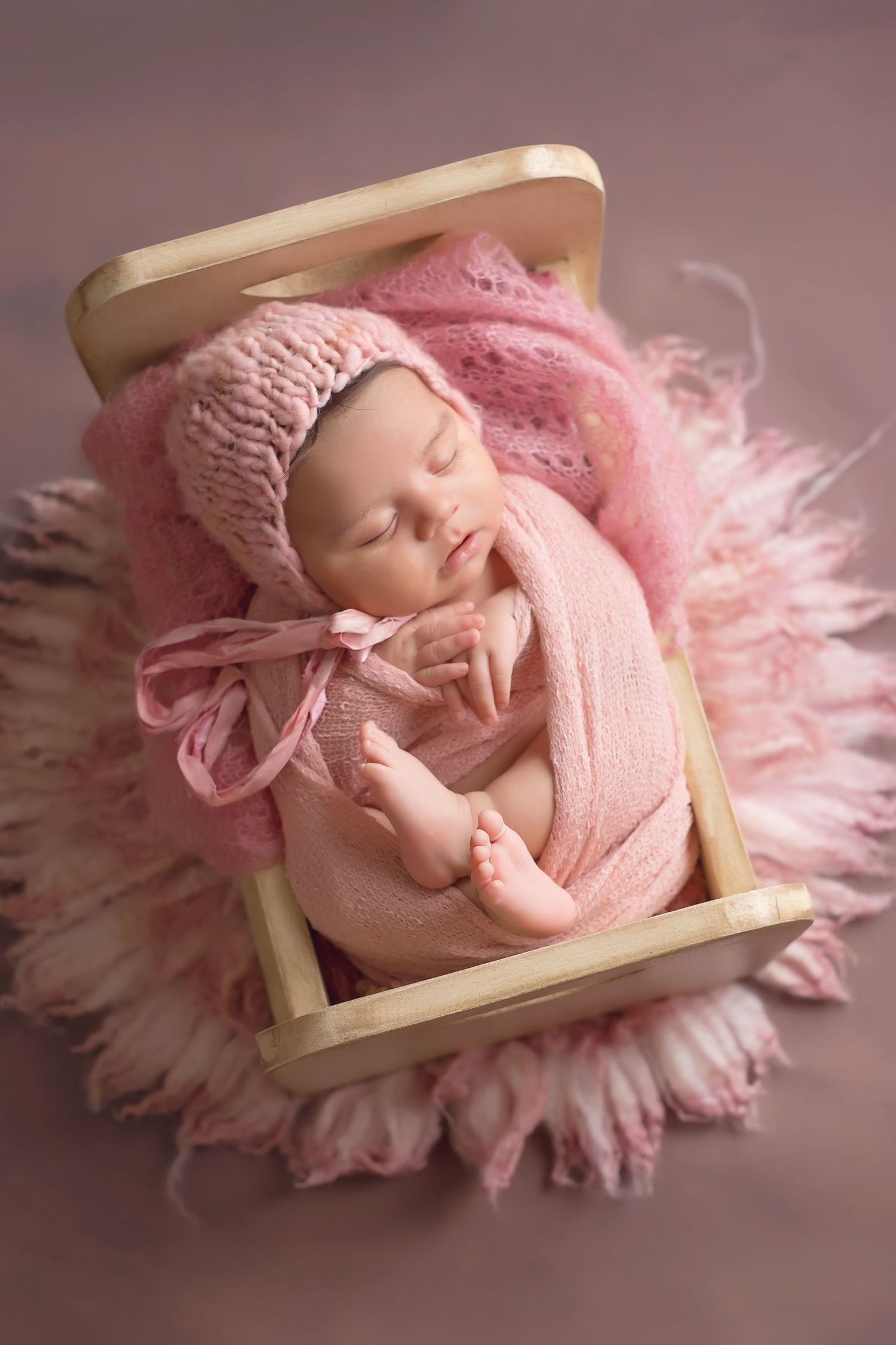 Sleeping baby wrapped in pink fabric and hat lying in wooden prop on pink fluffy background. San Diego Newborn Photographer