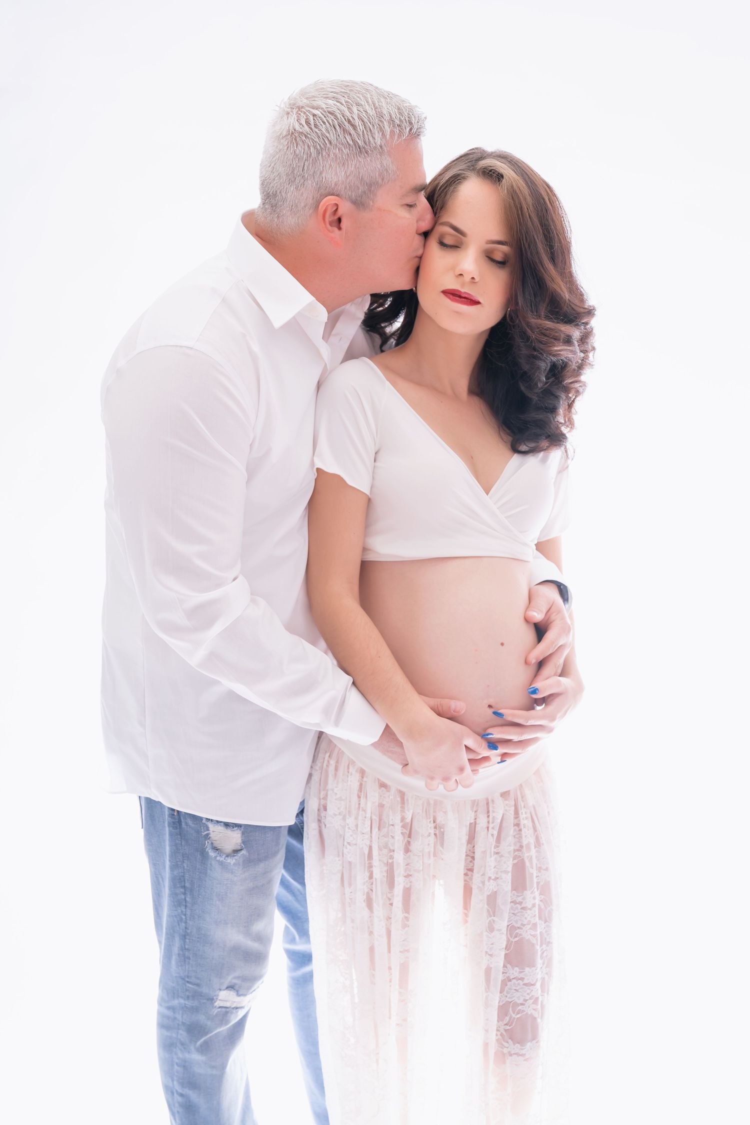 Intimate maternity portrait of couple in white clothing against light background.