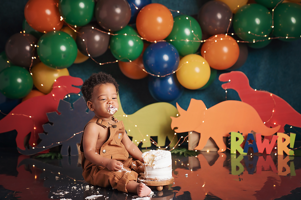 A baby boy smiles at his dinosaur-themed first birthday cake smash with colorful balloons and RAWR backdrop.
