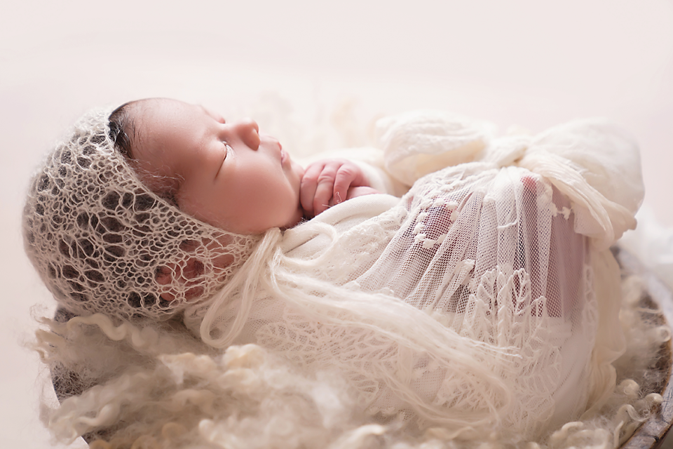 Newborn baby sleeping peacefully wrapped in cream lace fabric wearing a delicate knit bonnet in a soft white nest.