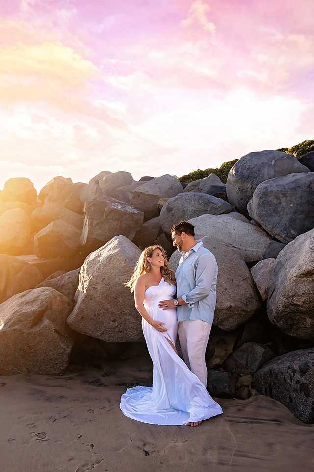 Pregnant couple in white attire embracing at sunset on a rocky beach, golden light glowing behind large boulders.