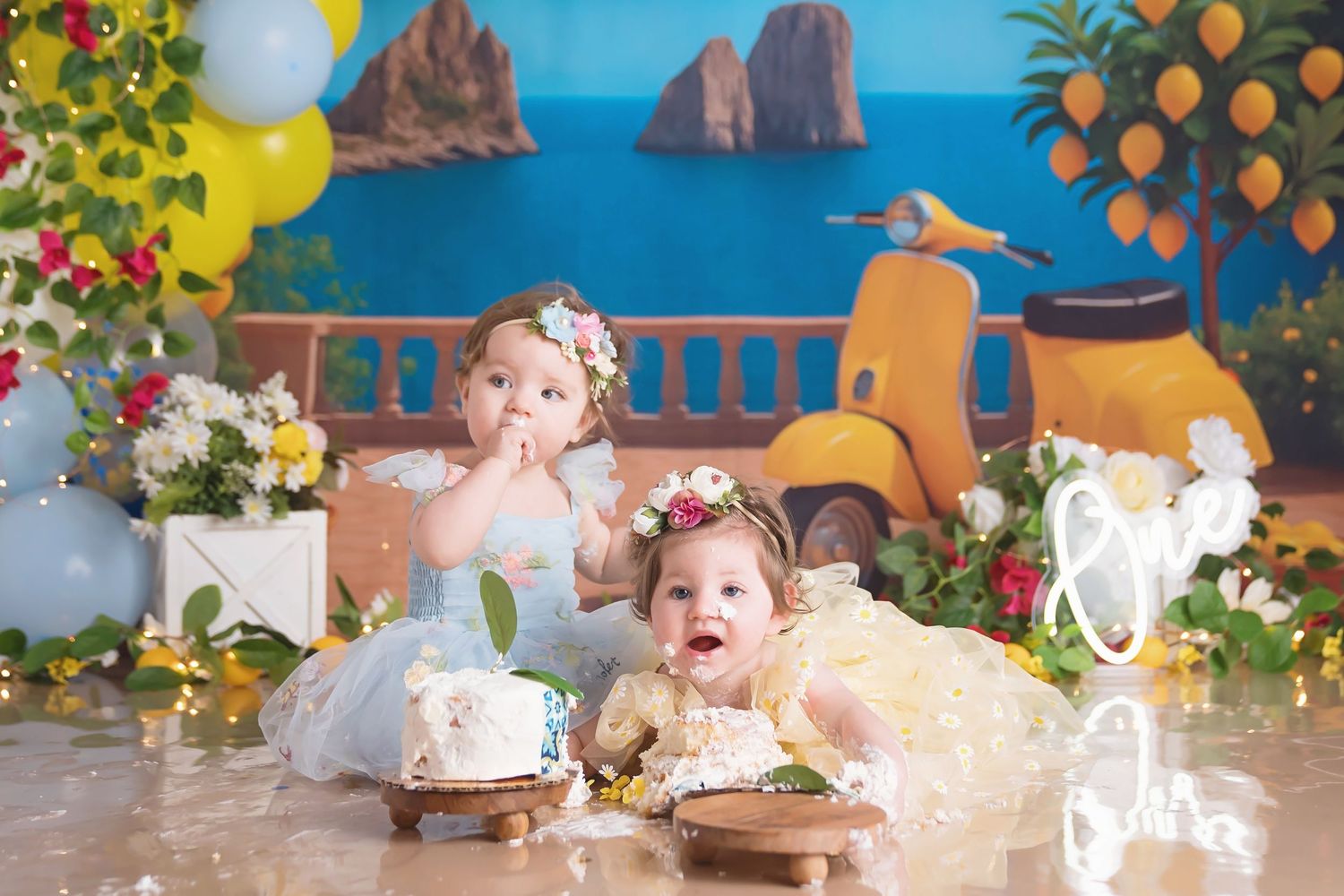 Twin baby girls in floral dresses celebrate their first birthday with smash cakes in a colorful Italian-themed photo set.