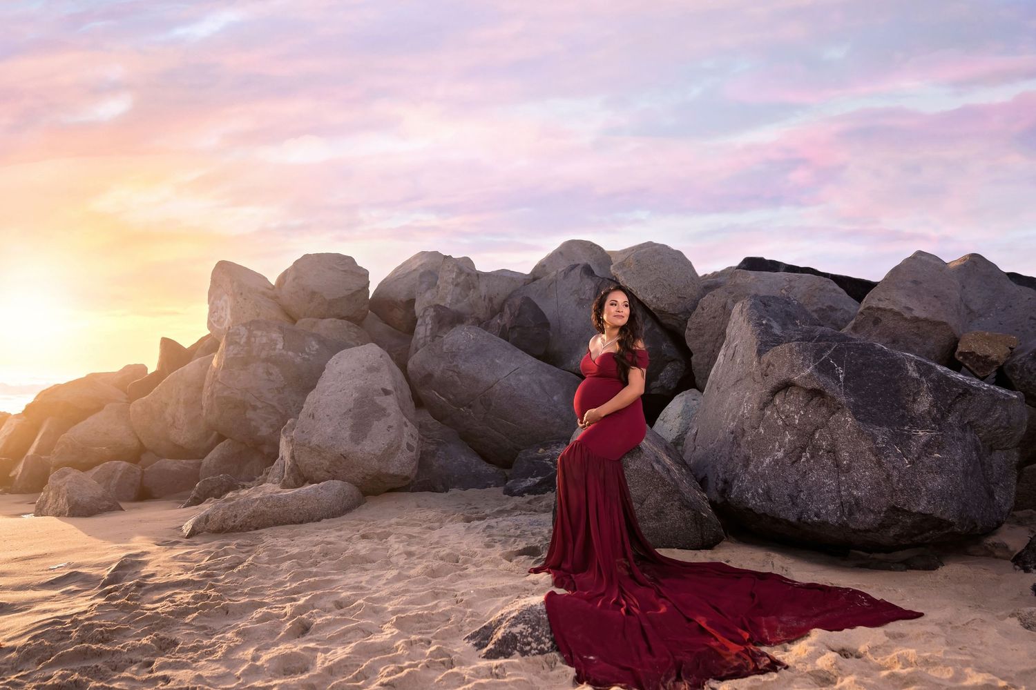 A dramatic red gown with long train flows against rocky coastal backdrop at sunset in san diego beach. San Diego maternity photographer