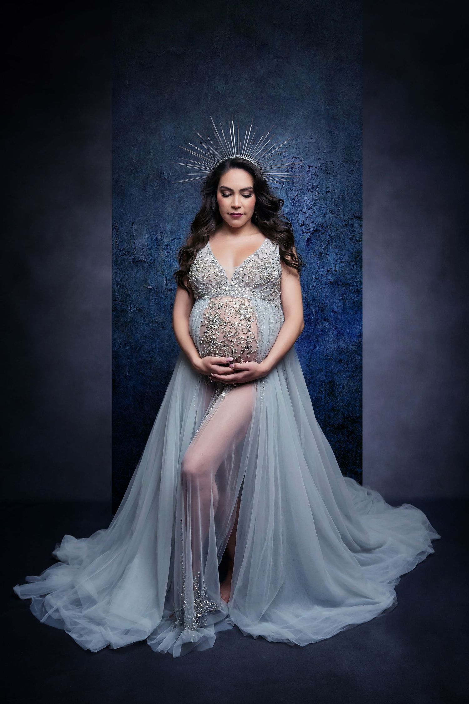 Ethereal maternity portrait with sheer silver gown and cape against blue backdrop