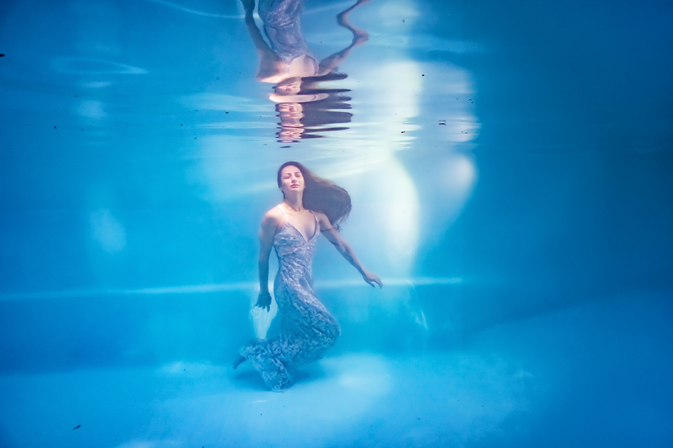 Woman in elegant gown swimming gracefully underwater in a blue pool, hair flowing, resembling a mermaid.
