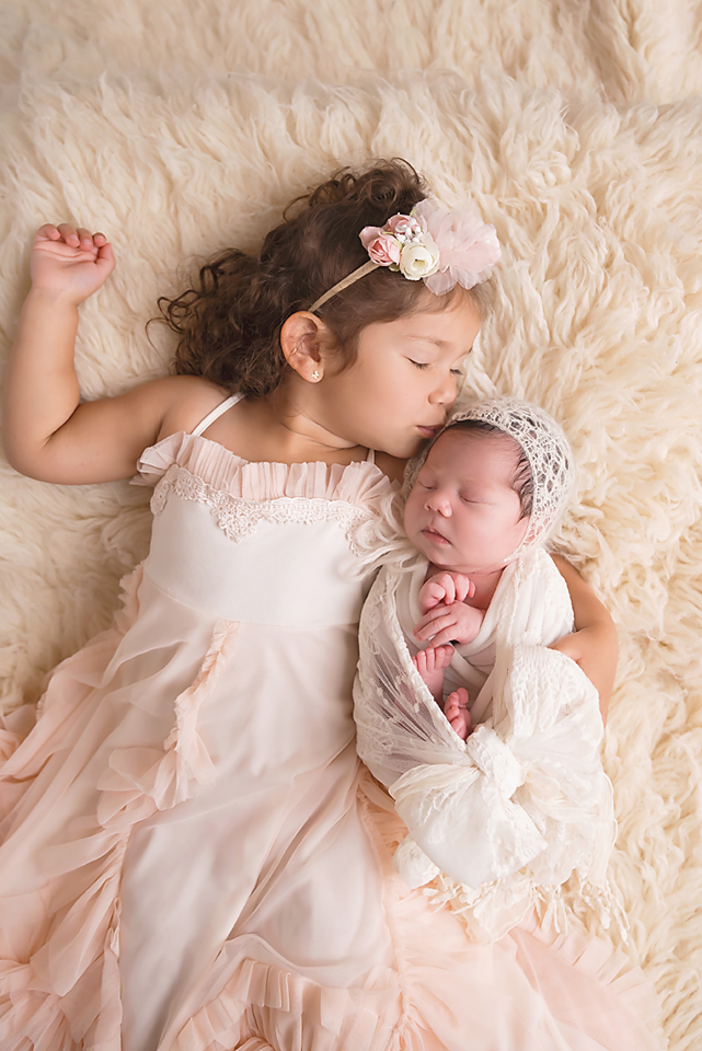 Young girl in pink dress kisses sleeping newborn sibling wrapped in white lace on a fluffy cream rug.