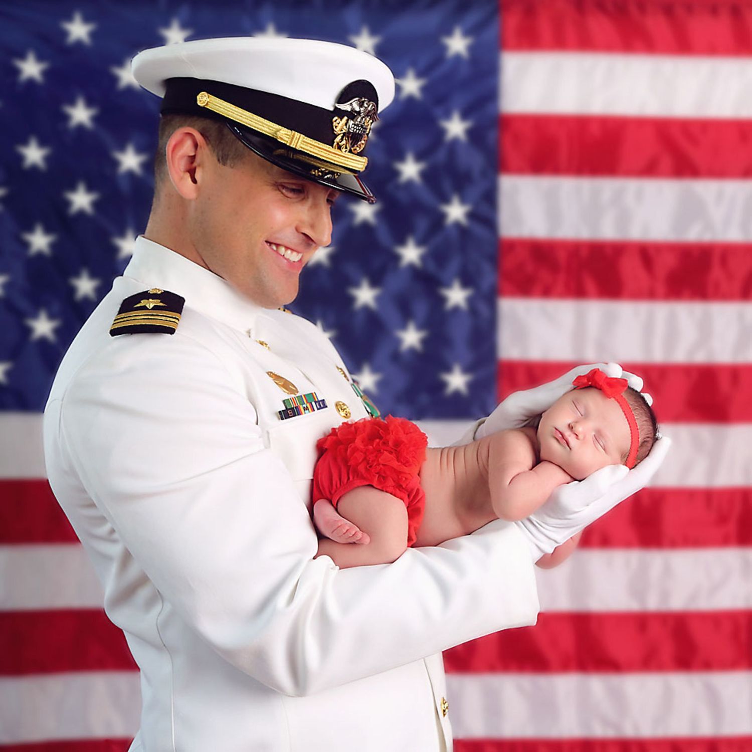 Military officer in white uniform poses with American flag backdrop with newborn baby in his hands. San Diego Newborn Photographer