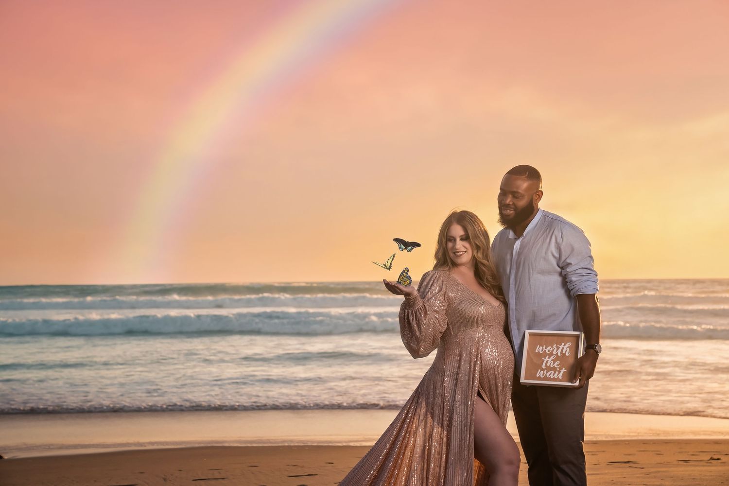 Pregnant couple poses on beach at sunset with rainbow, woman in sequin gown holding bird butterfly, man holding chalkboard sign.