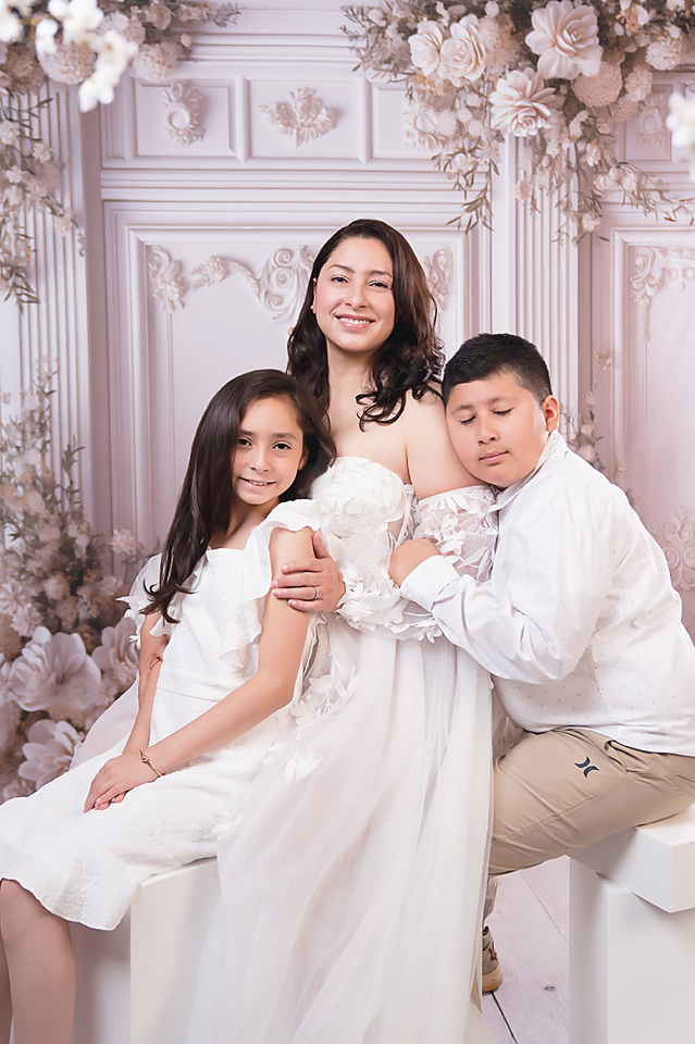 A pregnant woman in a white gown poses with two children in a floral studio setting.