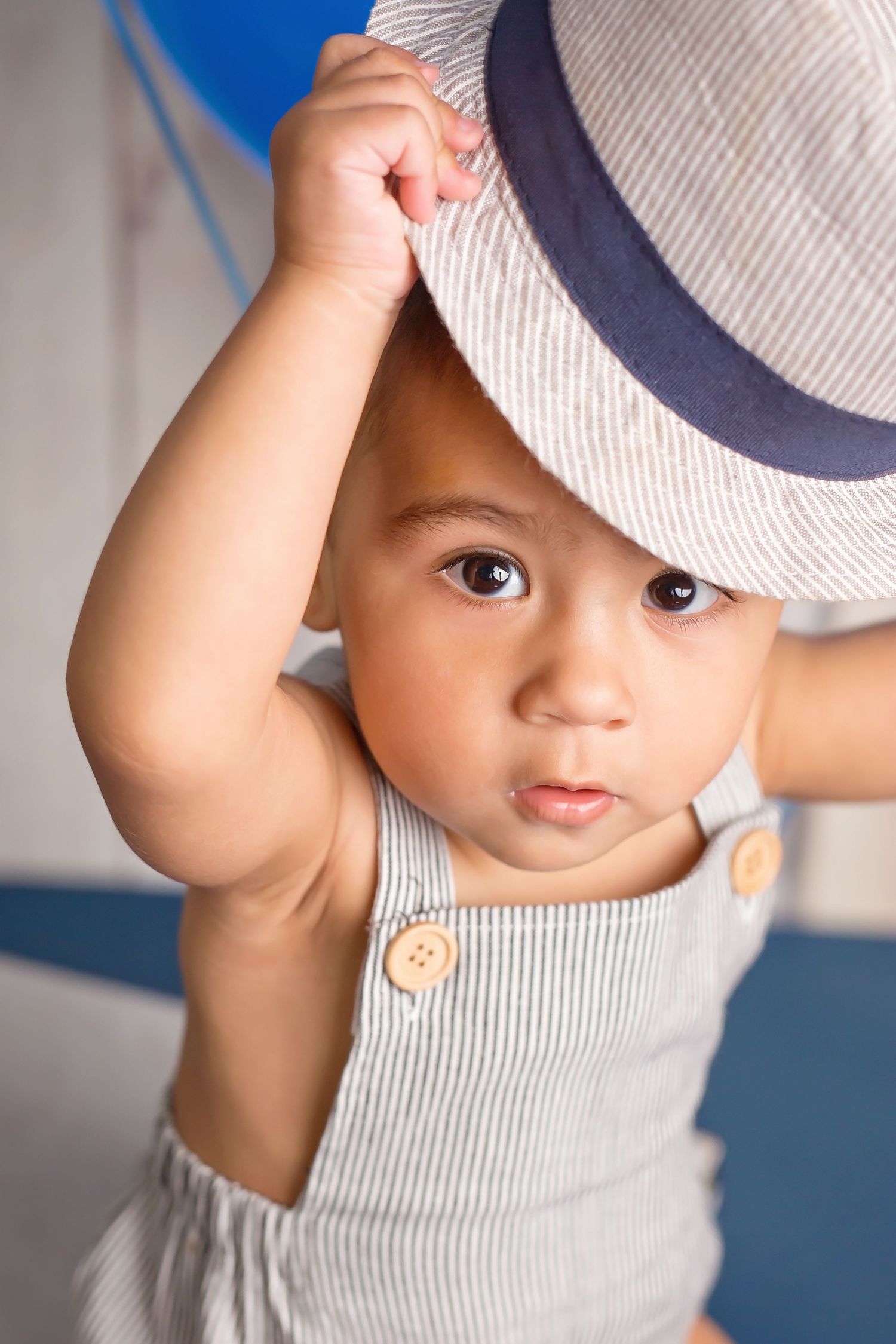Small child wearing a striped cap and light colored outfit peeks playfully at the camera. San Diego cake smash photographer