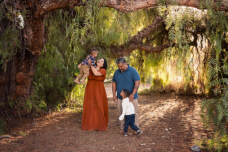 Family of four enjoying a sunny outdoor photo session under a large tree canopy on a dirt path.