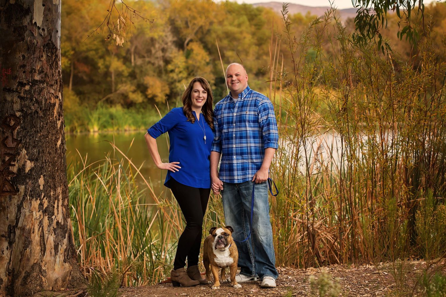 Couple in coordinating blue outfits posing beside a lake with autumn foliage