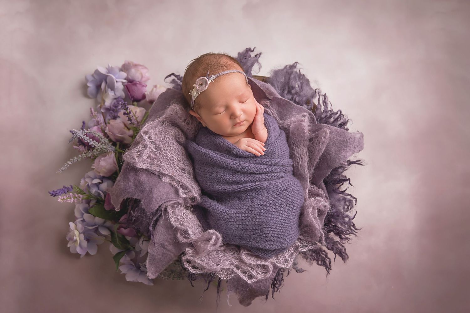 Newborn photography setup featuring purple wraps and delicate floral arrangements. San Diego Newborn Photographer