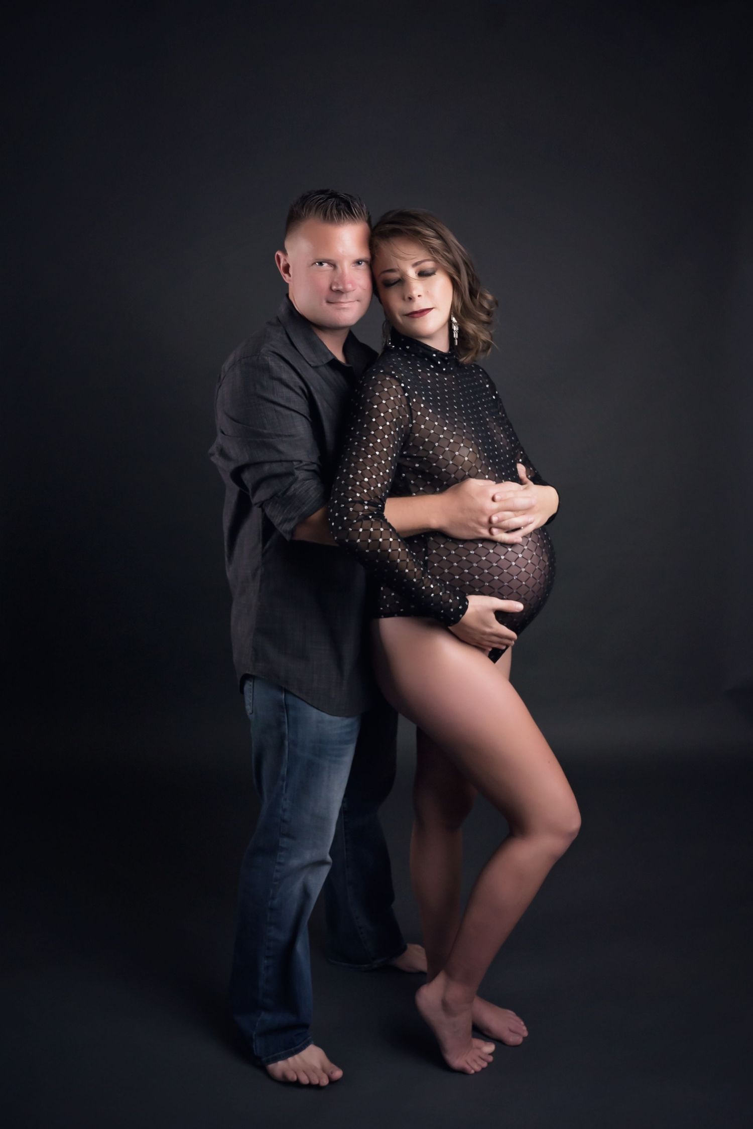 Intimate maternity portrait of couple in dark clothing against moody background.