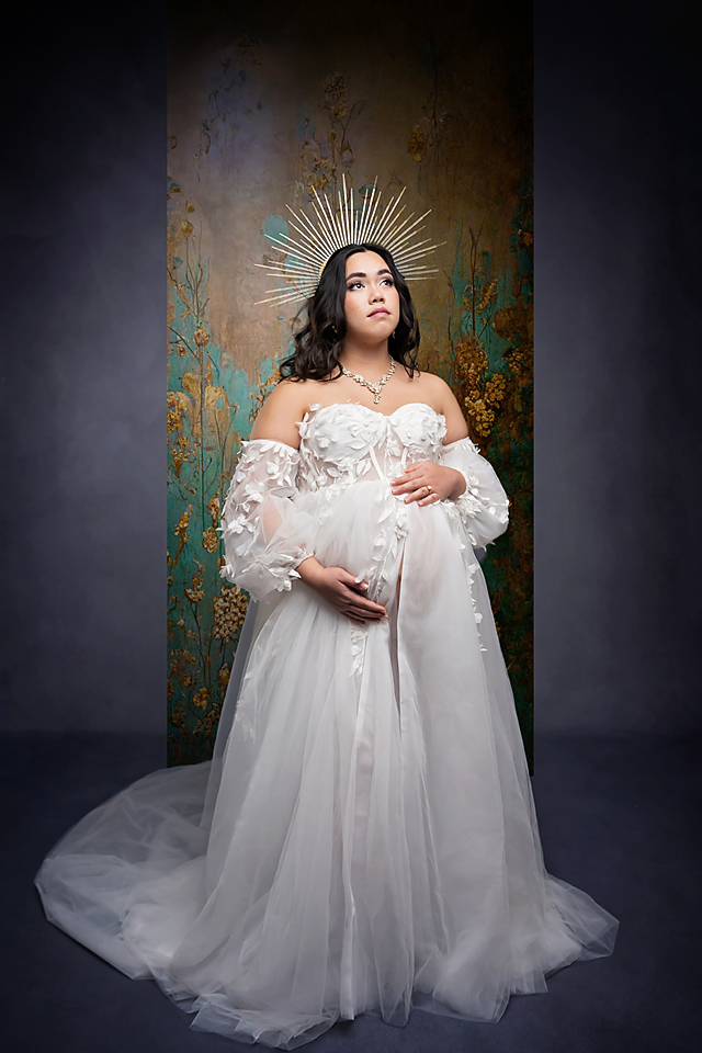 Pregnant woman in white tulle gown with floral corset and celestial halo crown poses gracefully in studio.