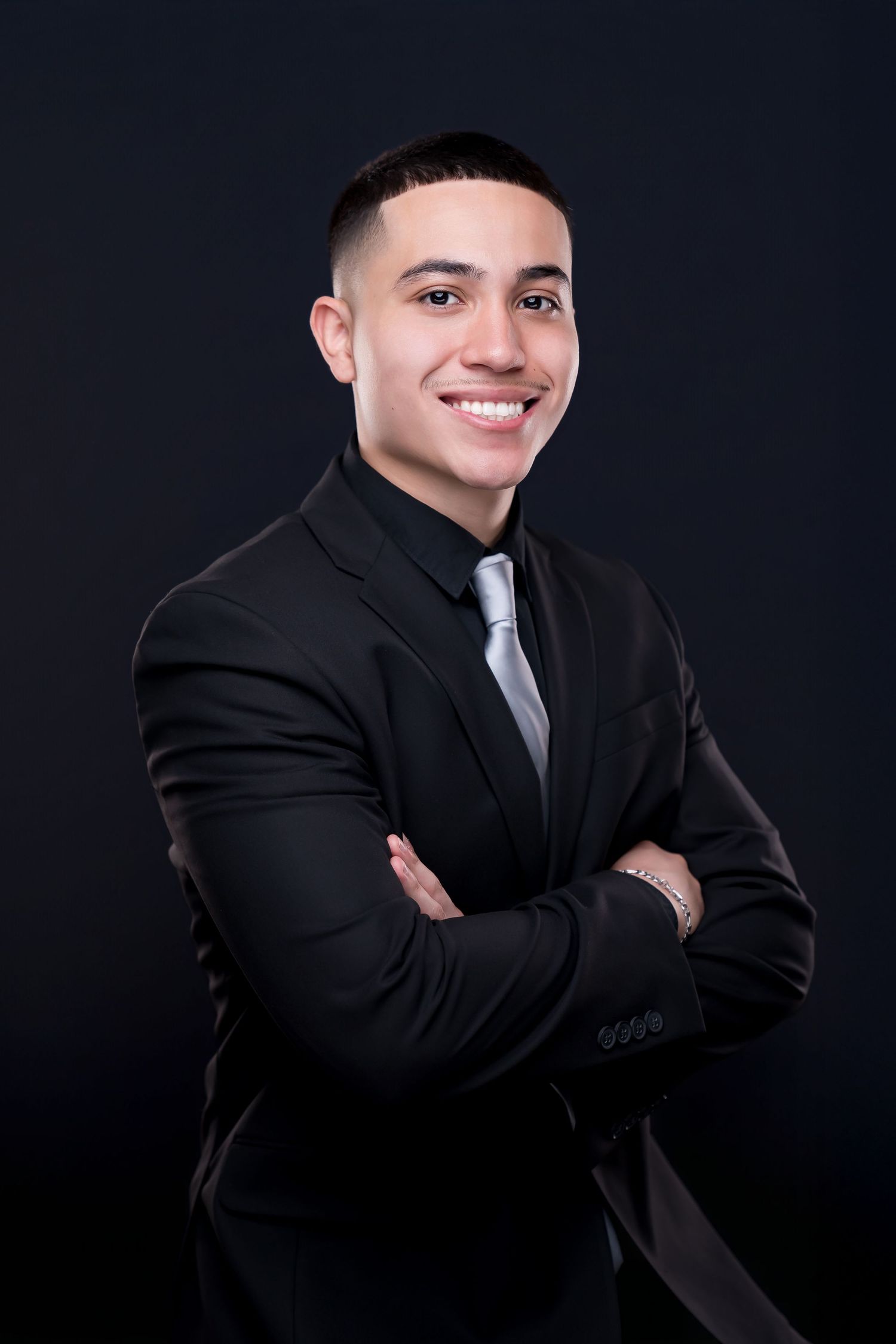 Young professional man in black suit and silver tie smiling with arms crossed against dark background.