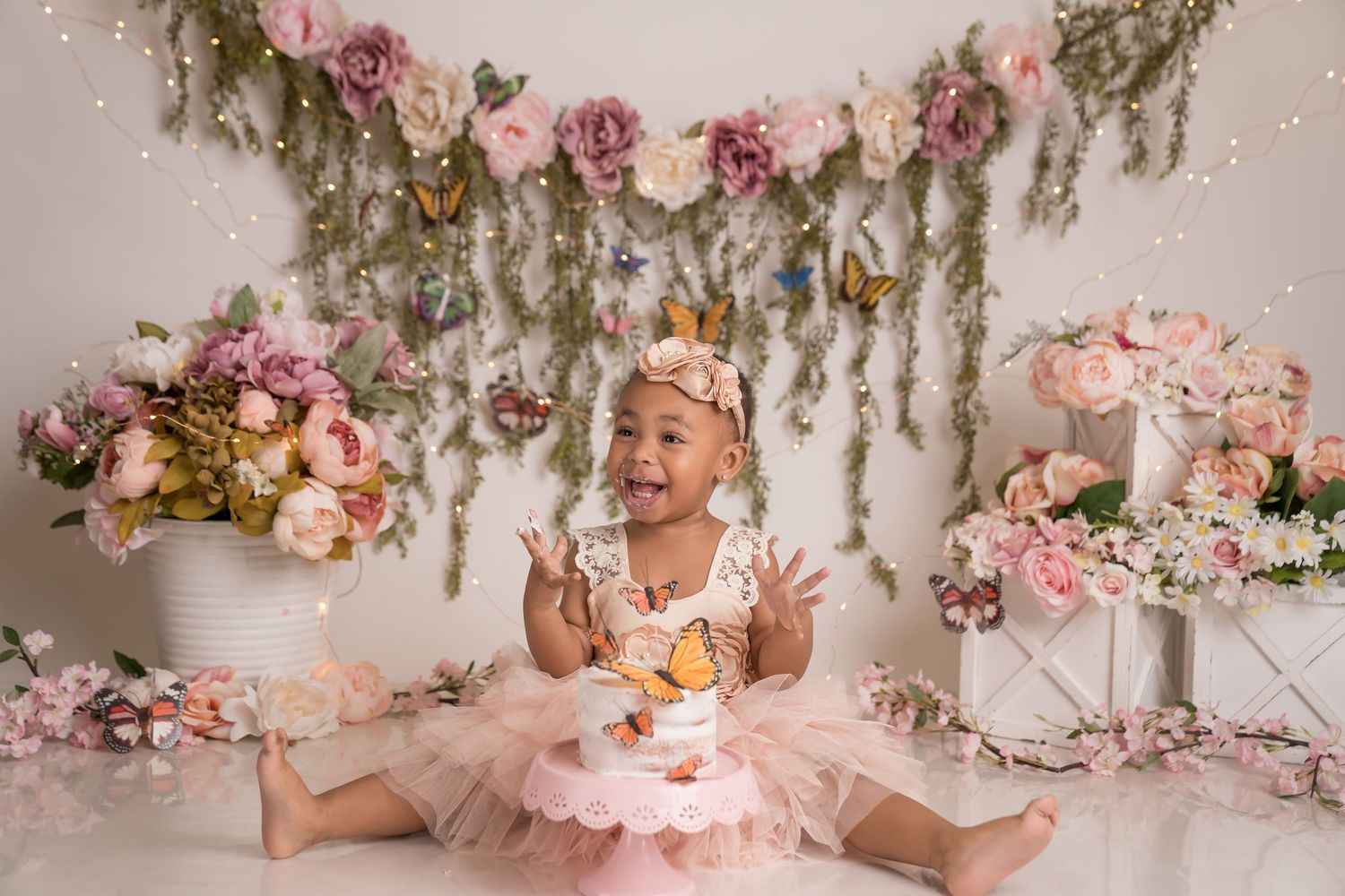 Whimsical floral cake smash photography setup with pink roses and greenery garlands against cream backdrop.