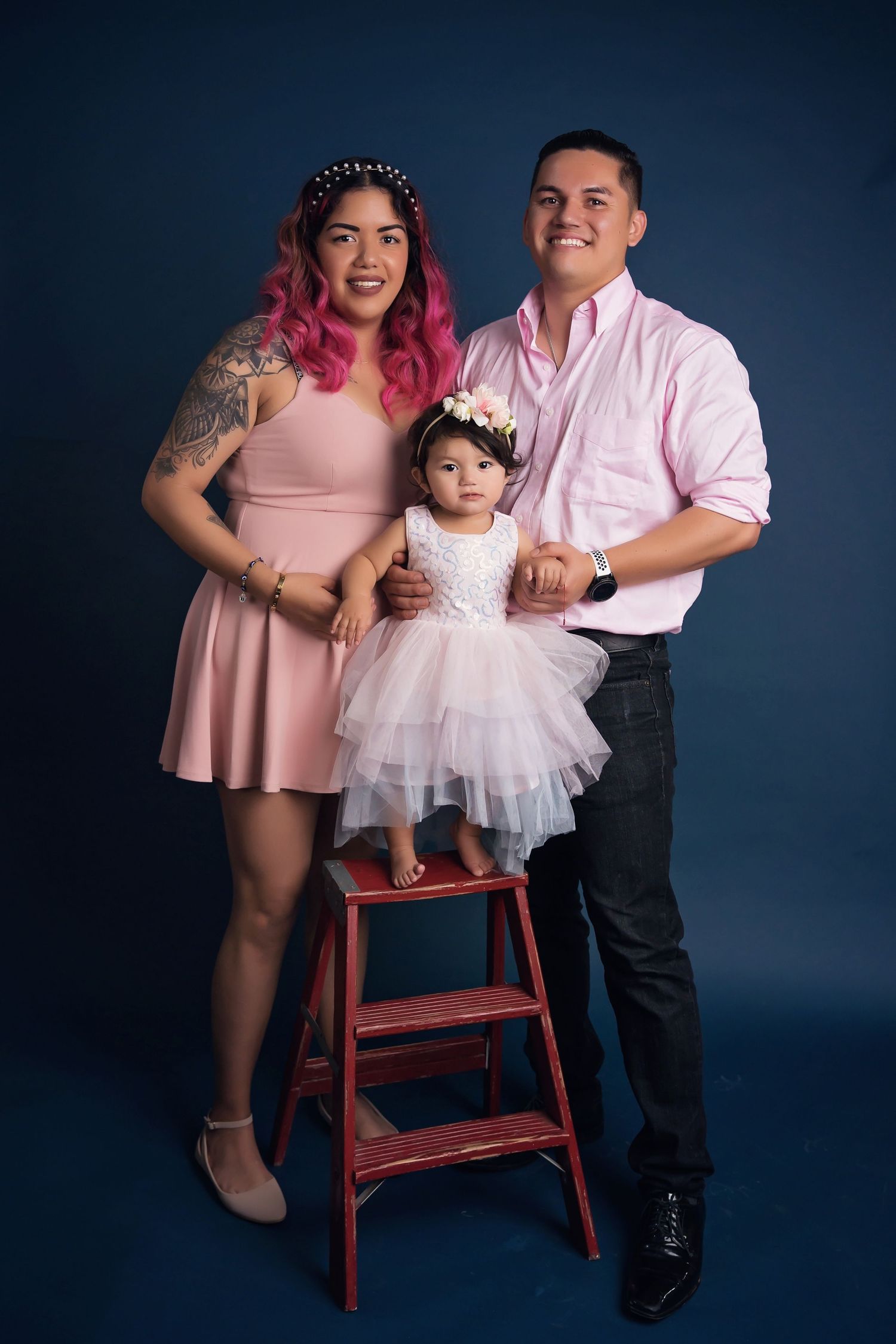 Family in pink coordinated outfits poses with red stepladder against navy studio background.