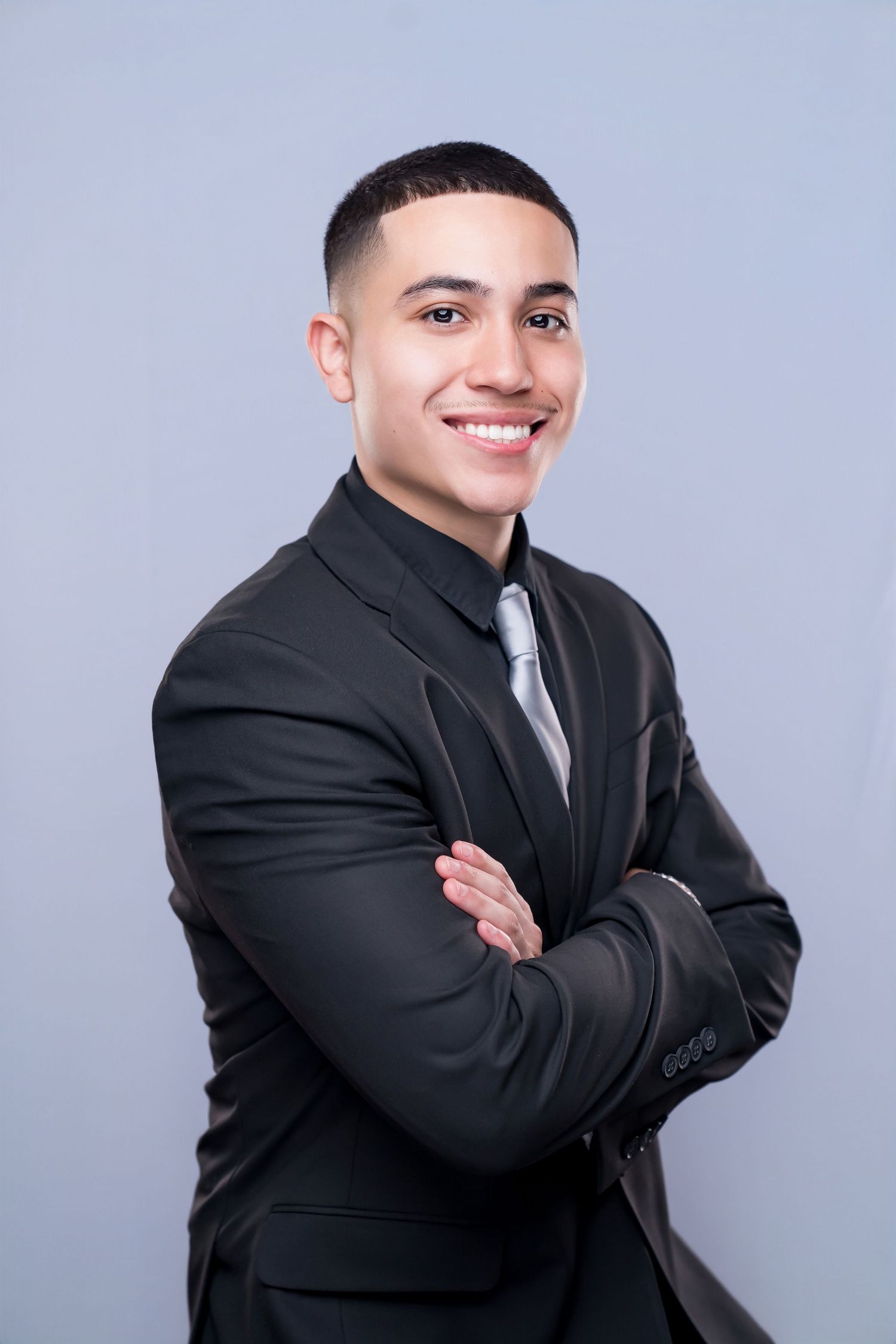 Young businessman in black suit and tie smiling with arms crossed against gray background.