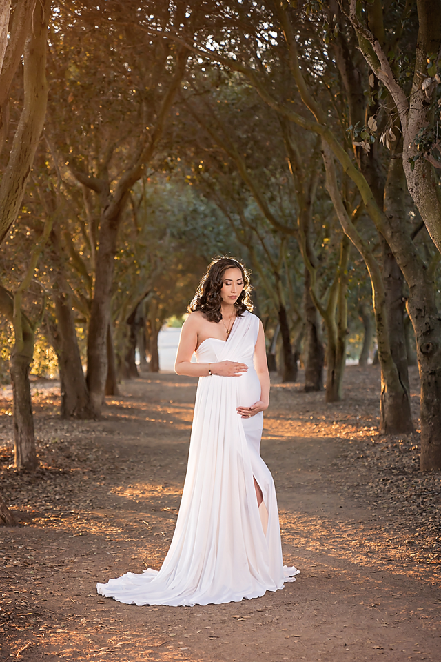 Pregnant woman in white one-shoulder gown poses gracefully on a tree-lined path during golden hour.