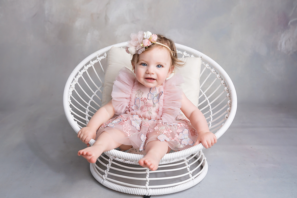 Smiling baby girl in pink lace romper and floral headband sitting in white wicker chair on gray background.