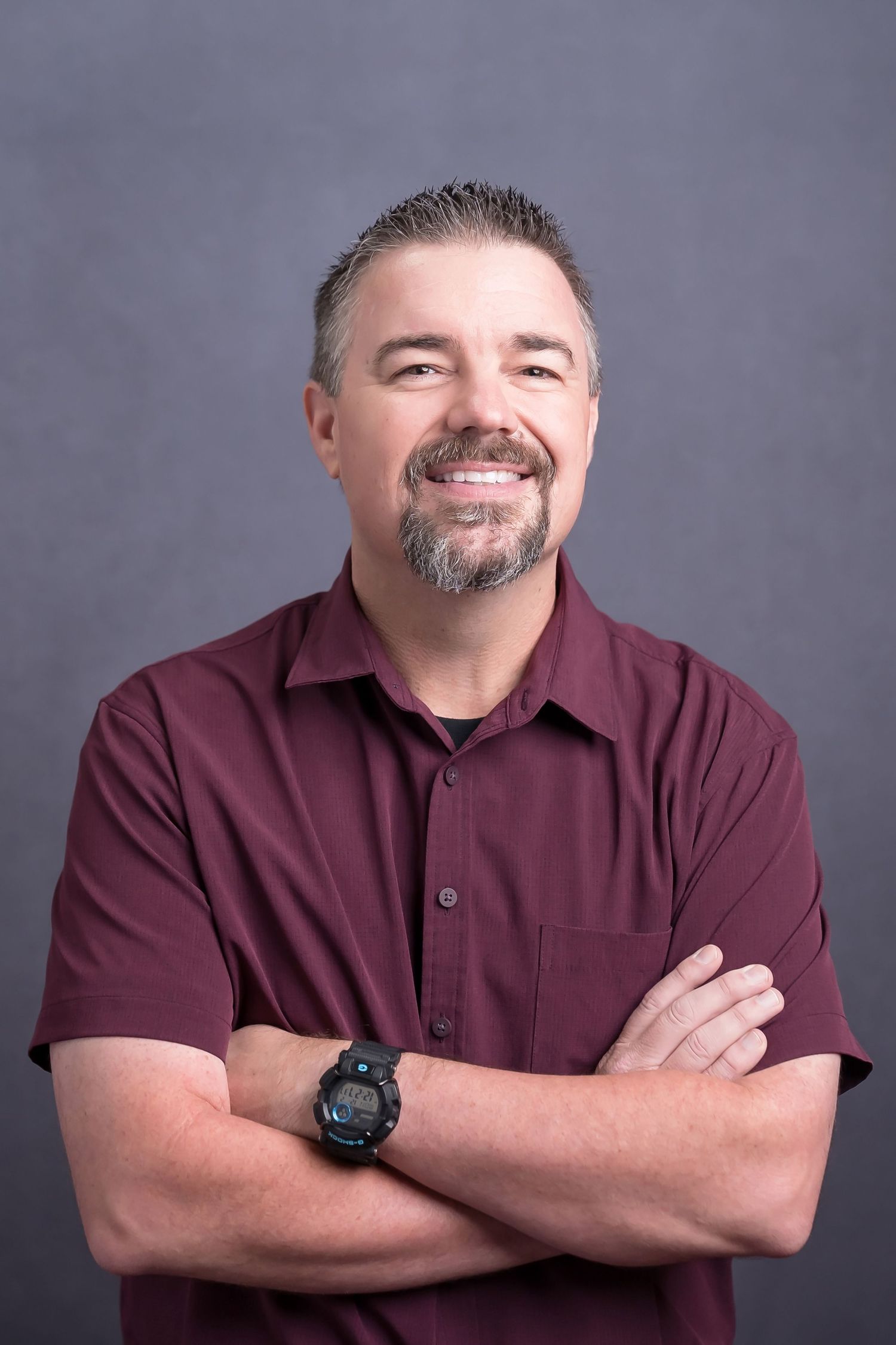 Man in burgundy button-up shirt posing with crossed arms against gray studio backdrop