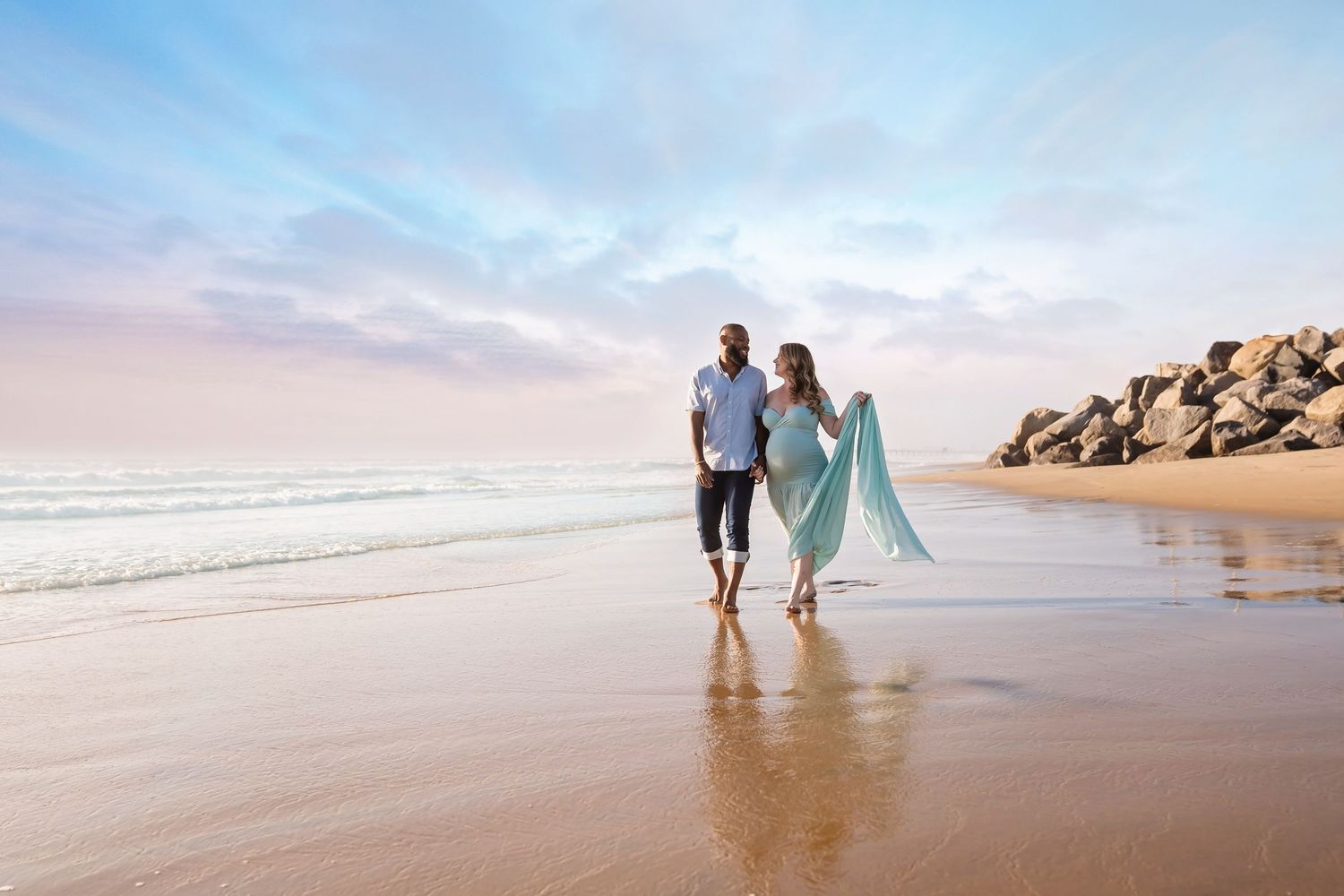 Couple walking along scenic beach at sunset with flowing fabric creating romantic silhouette against ocean waves. San diego beach maternity