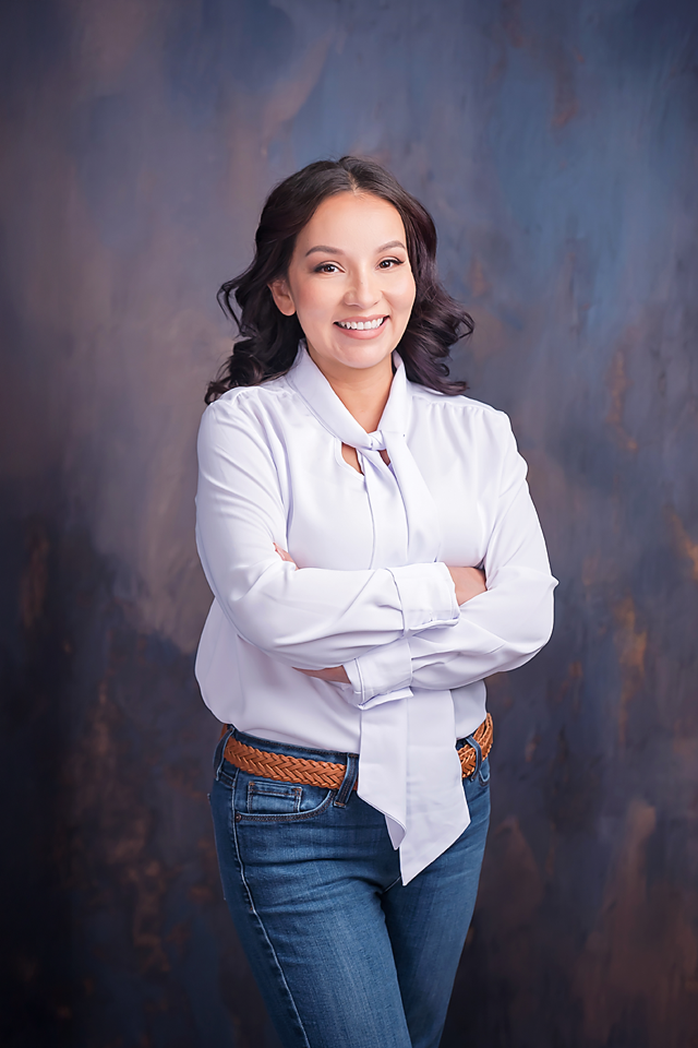Smiling woman in white blouse and jeans with arms crossed poses confidently against a dark textured studio background.