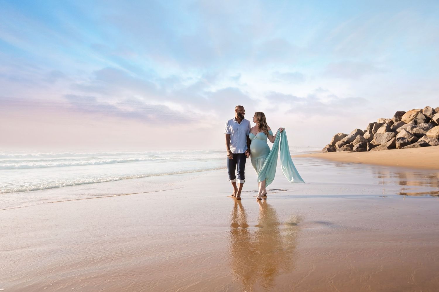 Maternity portrait of couple walking hand in hand on a sandy beach at sunset, with rocks and pastel sky in the background.