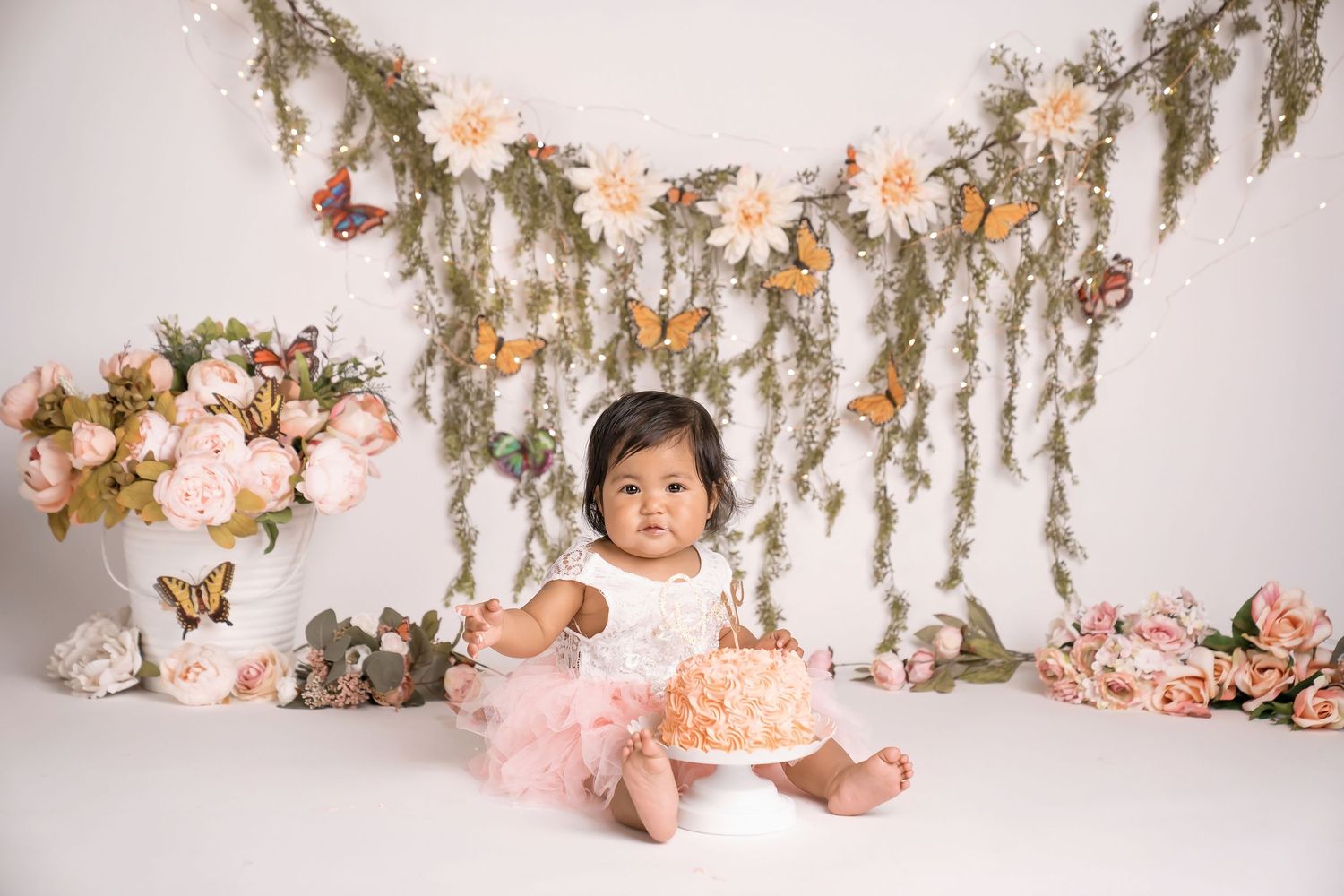 Baby girl in pink tutu sits with smash cake surrounded by flowers and butterflies for first birthday photo.